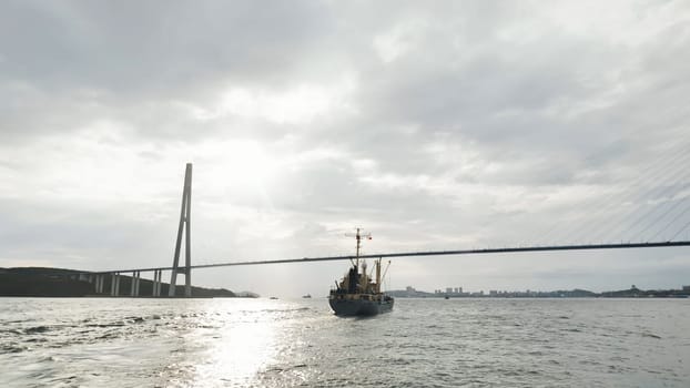 Cargo ship sailing under the russky bridge, vladivostok, russia, on a cloudy day with sun shining through the clouds