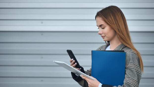 Professional woman working remotely, multitasking with digital devices near contemporary corporate exterior, wearing business attire