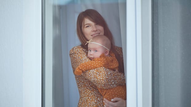 Mother and baby girl wearing matching floral print clothes are standing by window, embracing and smiling