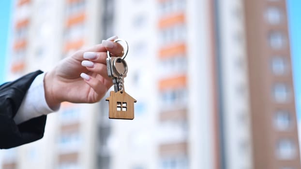 Realtor holding house shaped wooden keychain, standing near modern residential complex, representing property sale