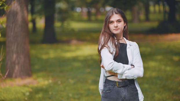Stylish young georgian woman with crossed arms stands confidently in a lush green park, enjoying the serene atmosphere