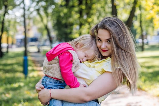 Mother cuddling peacefully sleeping daughter, sunlight filtering through verdant park landscape, tender maternal moment unfolding
