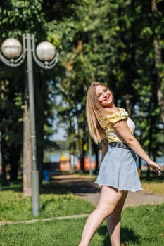 Blonde woman wearing a yellow top and denim skirt smiling and walking in a park during a sunny day