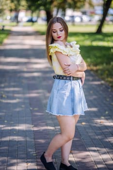 Fashionable young woman wearing a ruffled yellow top and denim skirt posing in a park during a sunny summer day