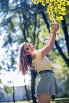 Teenager girl with long hair, wearing a yellow t shirt and a jeans skirt, is enjoying a sunny day in a park, touching the leaves of a branch
