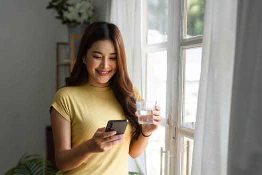 A young woman balances hydration and digital connection, smiling at her phone while enjoying a refreshing glass of water in a soothing home environment.