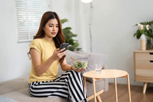 A woman sits on a sofa, checking her phone while having a nutritious salad, showcasing a balance of technology and wellness.