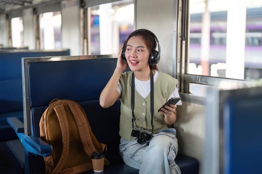 A smiling young woman joyfully interacts with her phone while wearing headphones on a train, capturing the essence of youthful enthusiasm during travel.
