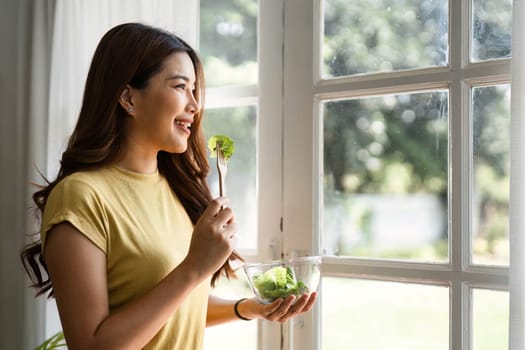 With a smile, a young woman enjoys her healthy salad, embracing the moment by the window and reflecting on the joy of mindful eating amidst a pleasant atmosphere.