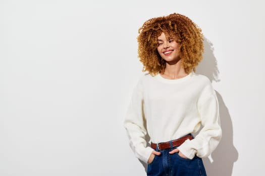 Smiling young woman with voluminous curly hair wearing a cozy white sweater stands confidently against a light background, exuding warmth and style Ideal for fashion and lifestyle content