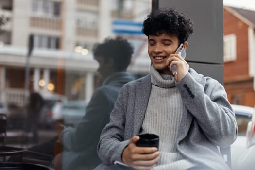 Young man talking on cell phone with cup of coffee in hand while sitting in front of window. fashionable and trendy concept.