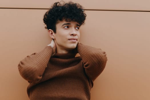 Fashion portrait of a young man with curly hair wearing a brown sweater, leaning against a wall with his hands on his head and looking at the camera.