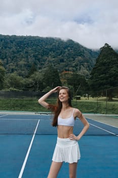 Happy young woman in a white sports outfit poses on a tennis court, showcasing a vibrant mountain backdrop Perfect for themes of health, fitness, and active lifestyles
