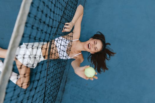 A joyful young woman in sporty attire, playfully holding a tennis ball, captures the essence of an active lifestyle and team spirit on a blue tennis court