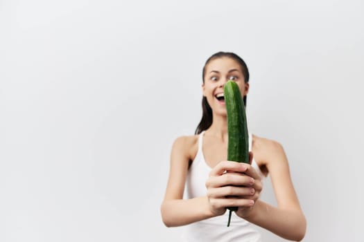 A cheerful woman joyfully holds a cucumber in her hands, wearing a simple white top against a clean light background Her expression conveys happiness and playfulness, emphasizing a fresh and healthy lifestyle