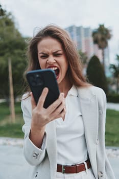 Frustrated businesswoman in a light suit passionately engaging with her smartphone in a chic outdoor location, reflecting the challenges of modern professional life.