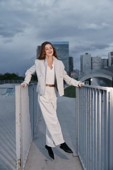 Confident woman in beige business suit walks on urban bridge under cloudy sky, smiling, showcasing modern fashion style and professional elegance in city environment