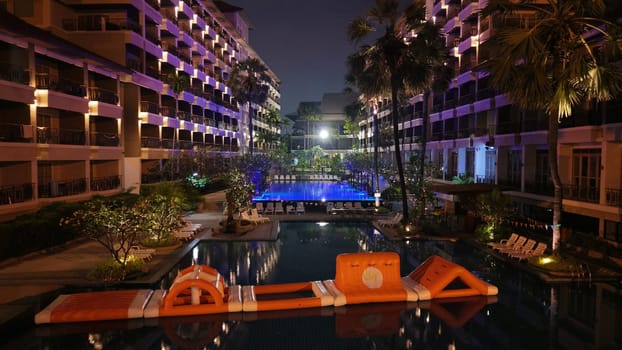 Orange inflatable obstacle course floating on a luxurious hotel pool at night, illuminated by the surrounding buildings and palm trees, creating a vibrant and inviting atmosphere