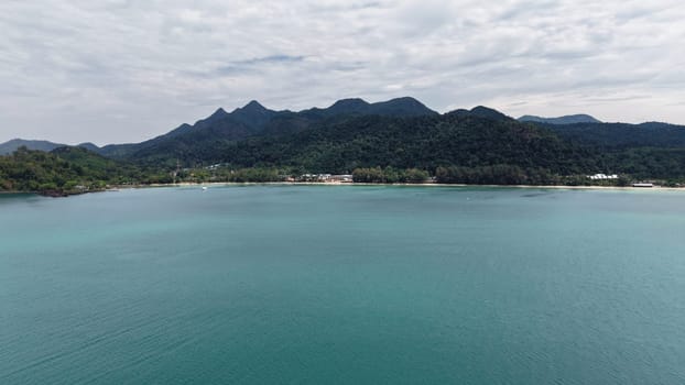 Turquoise water gently laps against the sandy shore of a tropical island, with lush green mountains rising majestically in the background under a cloudy sky, creating a serene and picturesque scene
