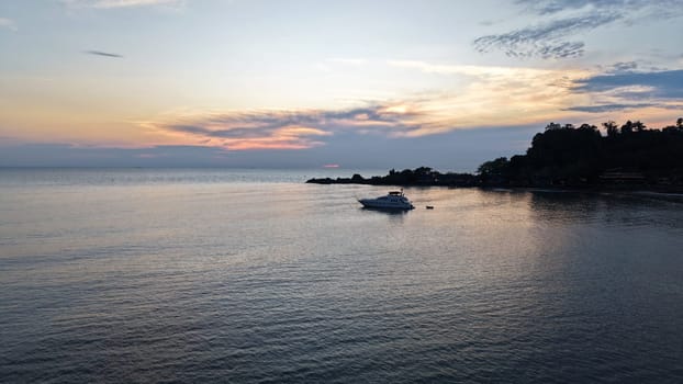 White luxury yacht rocking gently on calm ocean waters near a tropical island, with vibrant sunset hues of orange and pink reflecting on the tranquil sea