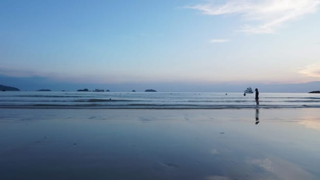 Woman standing in shallow water reflects on wet sand as sun sets over calm ocean with boats and islands in background, creating serene tropical paradise scene
