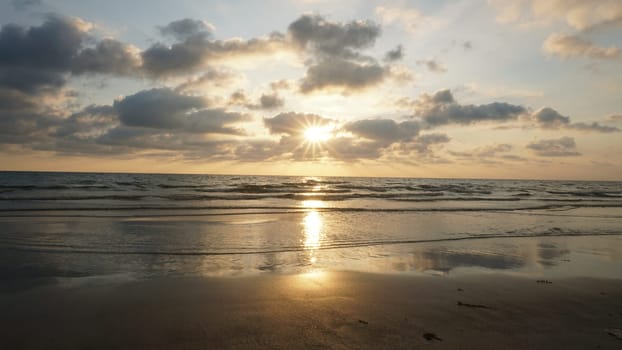 Soft sunlight filters through a cloudy sky, illuminating gentle waves rolling onto a wet sandy beach, creating a serene and reflective seascape at sunset