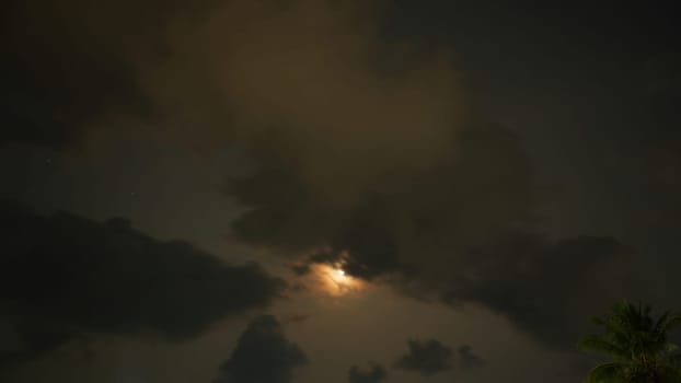 The moon shines through dark clouds at night, creating a moody and atmospheric scene with a few stars visible and a palm tree silhouette in the corner