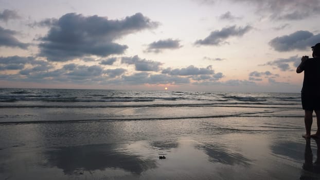 Photographer standing on a wet sandy beach at sunset, capturing vibrant sky colors reflected in gentle waves as the sun sets below the horizon