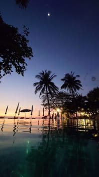 Palm trees swaying gently in the breeze as tourists relax in an infinity pool, taking in the breathtaking view of a vibrant sunset over a tropical ocean