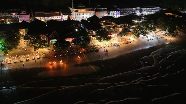 Aerial view of a vibrant beach town at night, featuring tourists dining at illuminated beachside restaurants and hotels glowing warmly along the shore
