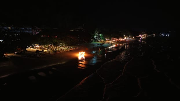 Tourists are enjoying a night party with a bonfire on the beach, creating a vibrant and festive atmosphere along the shoreline under the dark night sky