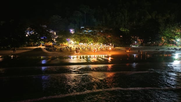 Tourists are enjoying a night party with a bonfire on the beach, creating a vibrant and festive atmosphere along the shoreline under the dark night sky