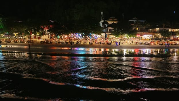 Lively night scene on a tropical beach with tourists dining and enjoying the vibrant atmosphere, colorful lights reflecting in the gentle ocean waves creating a magical ambiance
