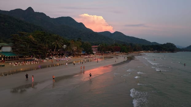 Tourists are walking along the beach at sunset, with restaurants and bars lining the shore, a mountain view in the background, and the sky reflecting on the wet sand in koh chang, thailand