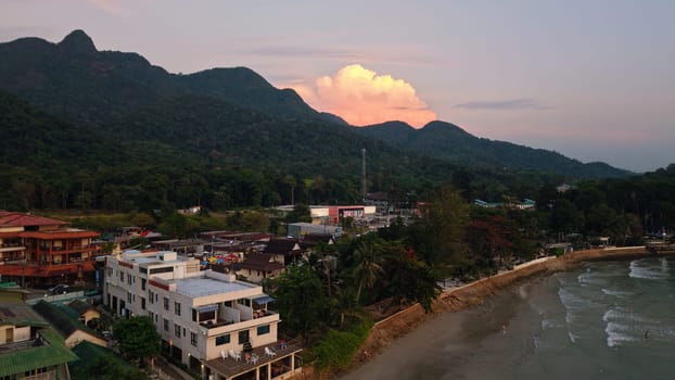 Tranquil coastal town nestled amidst lush green mountains at sunset, featuring a dramatic cloudscape illuminated by warm hues, creating a serene and picturesque scene