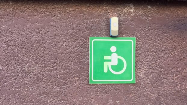 Close up of green wheelchair accessibility sign with doorbell on rough brown wall, minimal urban element representing inclusive access and emergency assistance for disabled individuals