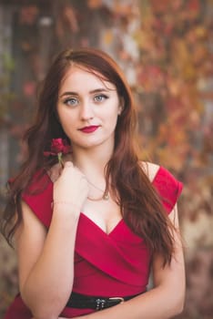 portrait of a young girl in a red dress against the background of autumn trees with a rose in her hands. thoughtful and romantic girl with long dark hair. emphasis on the red dress, lips, rose