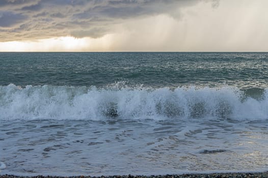 Waves roll onto a pebbled beach as dark clouds gather overhead during sunset. The ocean reflects light while the storm approaches, creating a captivating atmosphere.
