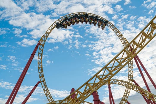 A thrilling roller coaster twists and turns against a backdrop of fluffy clouds and blue sky. Riders scream with excitement as the ride reaches its peak, creating a moment of pure exhilaration.