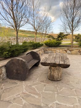 A peaceful outdoor space features rustic wooden seating and a table made from tree trunk materials, surrounded by manicured bushes and trees on a clear day.