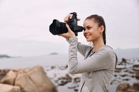 A young woman stands on a rocky beach, captured in a moment of joy while taking photos She wears a casual grey outfit, reflecting a blend of creativity and nature s beauty