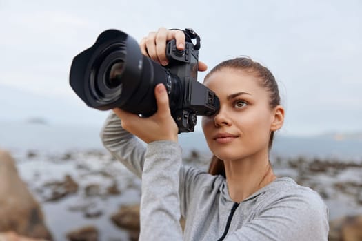 A young woman with long hair and a casual gray outfit focuses on her DSLR camera by the ocean, highlighting creativity and love for photography by the sea