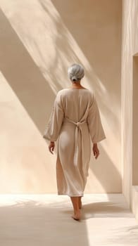 A senior silver-haired woman moves gracefully through a softly lit spa hallway, her robe flowing gently as she brushes her hands against a textured wall. The ambiance is calm and inviting.