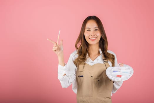 Asian beautiful young woman artist holding brush and paint palette, Happy female painting using paintbrush and palette with colors, studio shot isolated on pink background, Paintings and art equipment