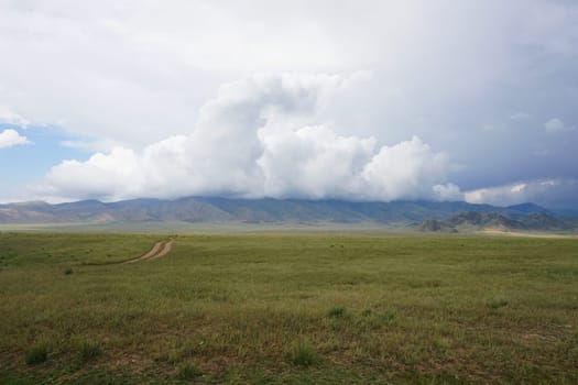 Vast steppe landscape in mongolia showcasing a winding dirt road, distant mountains rising majestically, and a dramatic cloudy sky enhancing the serene atmosphere of the wilderness