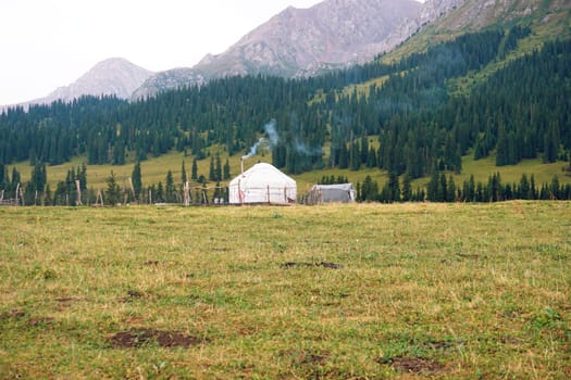 Smoke rising from the chimney of a yurt creates a warm atmosphere, surrounded by lush green hills and majestic mountains in a tranquil, nomadic landscape