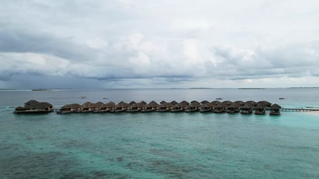 Picturesque view of overwater bungalows extending from a tropical island into the clear turquoise ocean, under a dramatic cloudy sky, creating a serene and luxurious vacation atmosphere