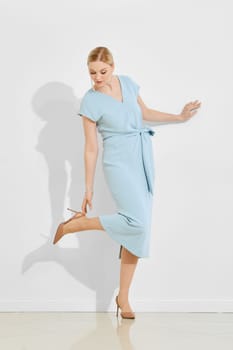 A woman is gracefully posing in a light blue dress, standing against a plain white backdrop.