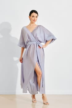 A woman showcases a stylish light lavender dress featuring a deep slit and relaxed sleeves. She stands confidently in front of a plain white backdrop, highlighting her fashion choice.