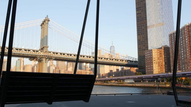 New York City, United States - 7 Sept 2023: Manhattan Bridge to Brooklyn. Pier 35 swings bench, waterfront downtown skyline cityscape. World Trade Center skyscraper, financial district morning sunset.